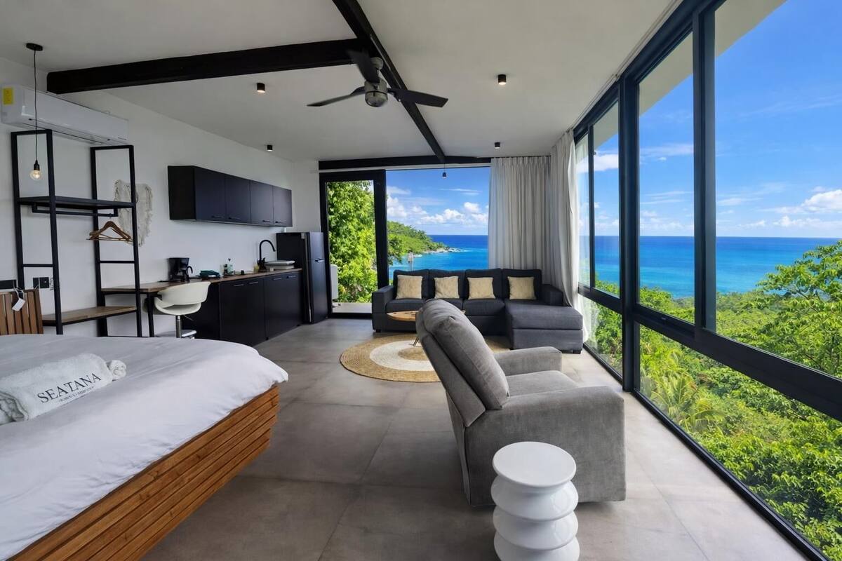 A modern bedroom with a bed, a desk, a sofa, and a view of the ocean.