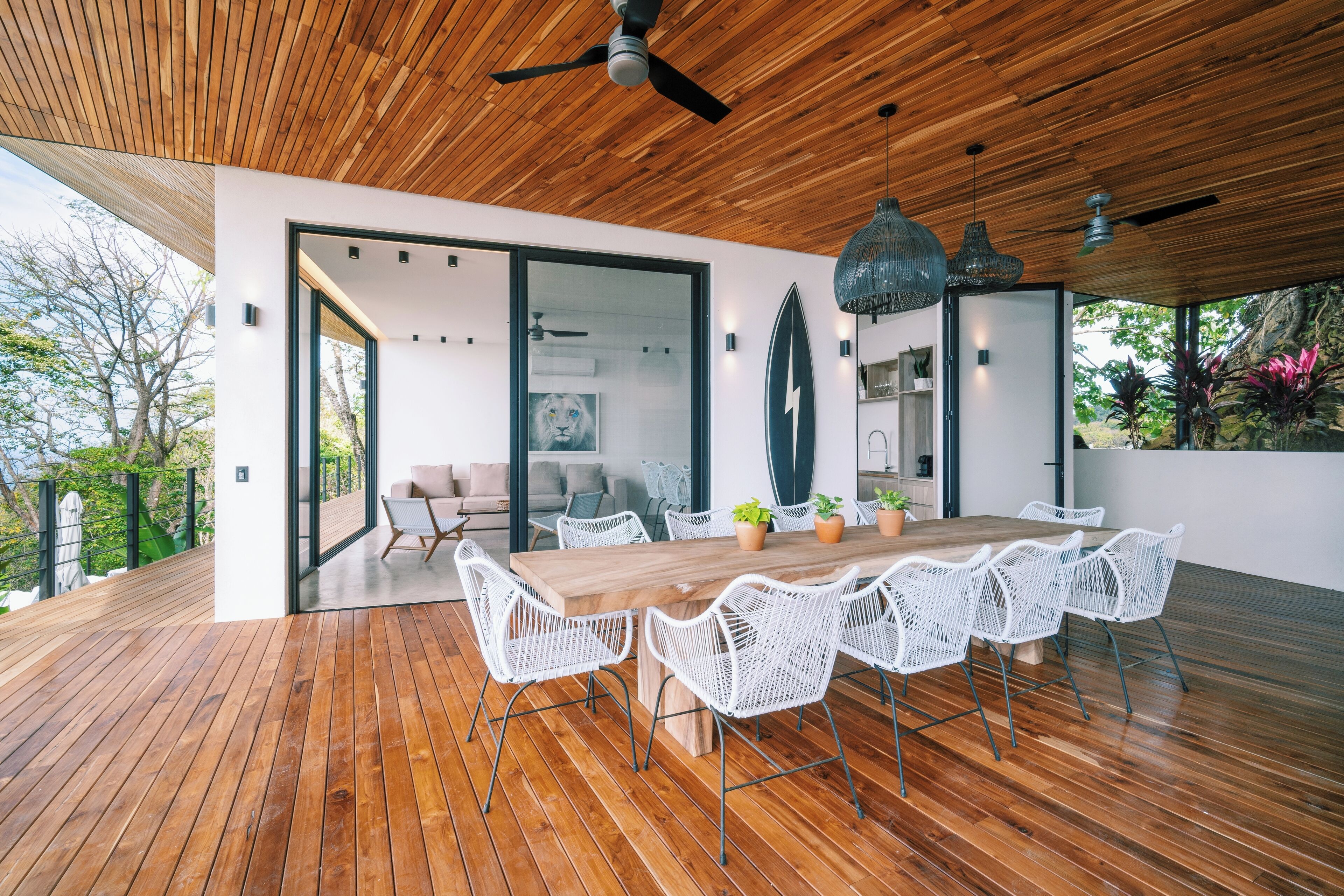 A modern dining area with a wooden table and white wicker chairs, a ceiling fan, and a view of the outdoors.