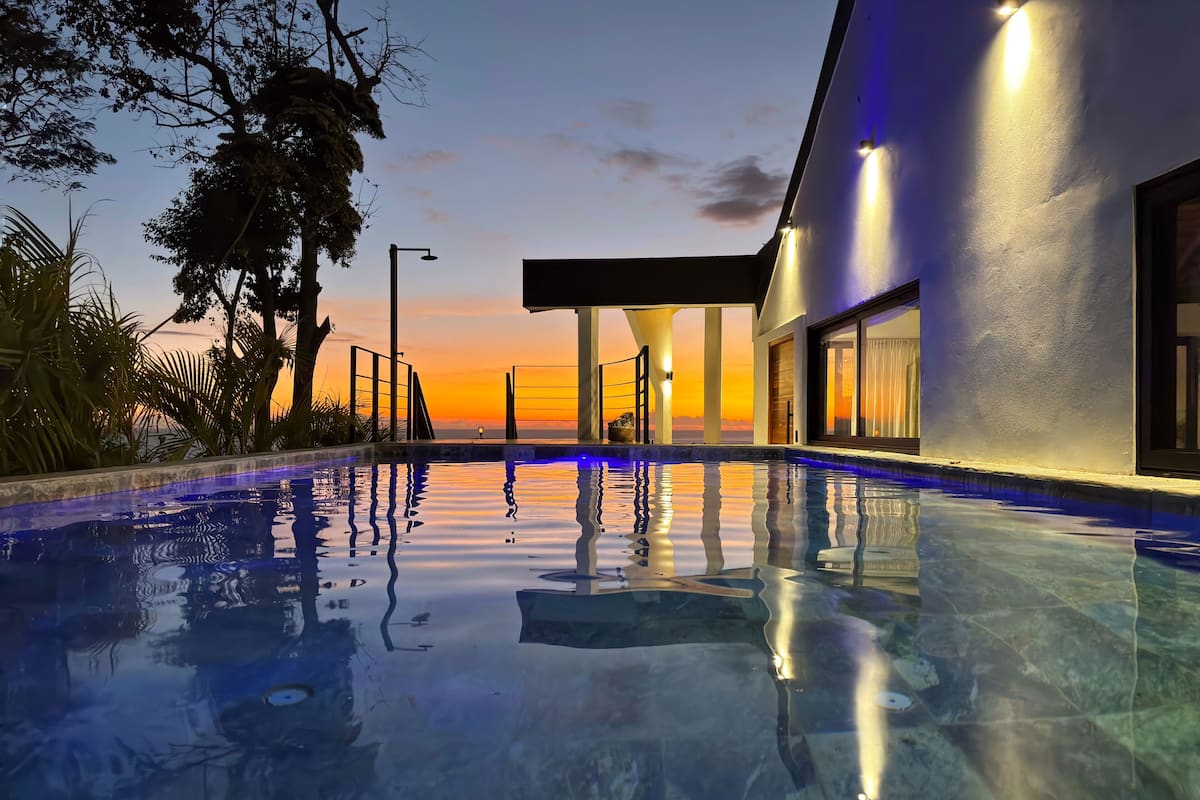 A modern pool area with clear water reflecting the sky, surrounded by lush greenery and a building with large windows.