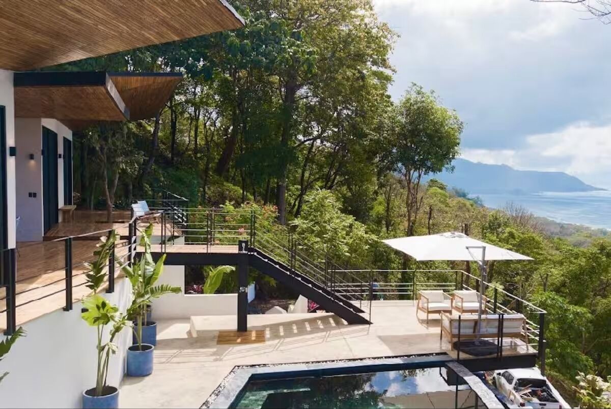 A modern outdoor pool area with a staircase leading to a deck, surrounded by lush greenery and overlooking a scenic view.