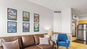 A living room with a brown leather sofa, a blue armchair, and a coffee table. There are four framed posters on the wall.