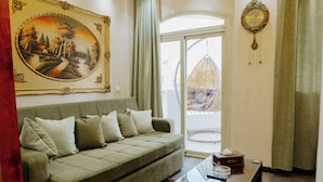Un salon avec un canapé vert, une table basse en verre, un tableau, une horloge et une vue sur l’extérieur, visible à travers une porte vitrée.