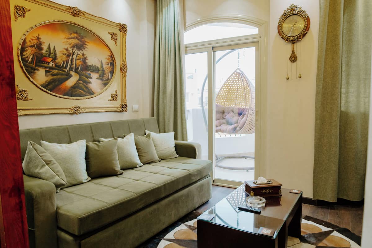 Ein Wohnzimmer mit einem grünen Sofa, einem Glastisch, einem Gemälde, einer Uhr und Blick durch eine Glastür.
