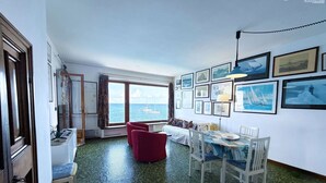 A room with a large window offering a view of the sea, a dining area with a table and chairs, and a bed with a view.