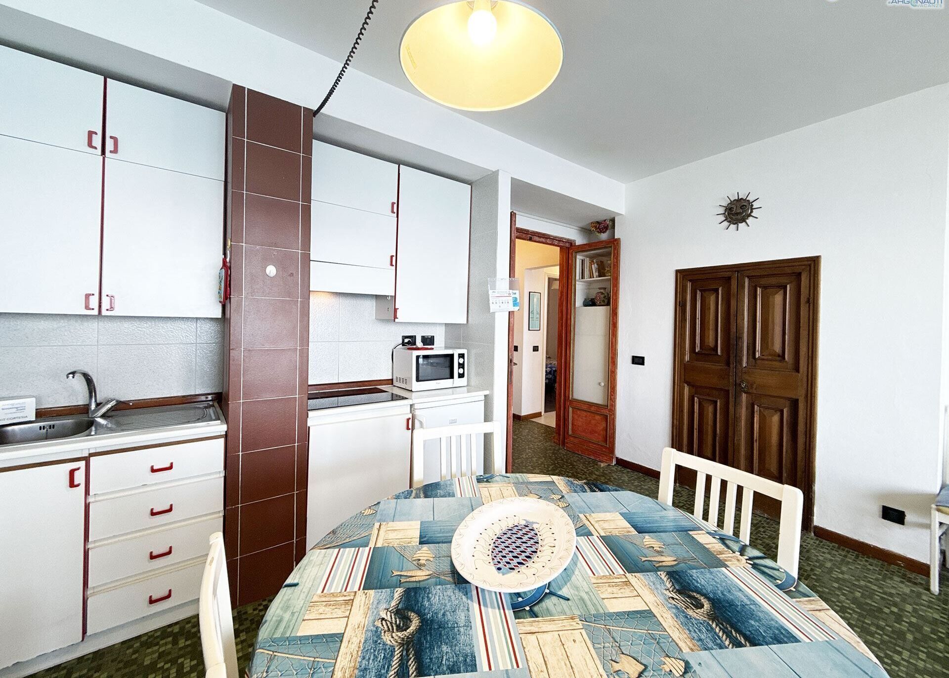 A compact kitchen with white cabinets, a microwave, and a sink. There's a dining table with a patterned tablecloth and a chair.