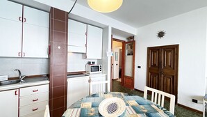 A compact kitchen with white cabinets, a microwave, and a sink. There's a dining table with a patterned tablecloth and a chair.