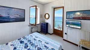 A bedroom with a bed, a window with a view of the sea, a door leading to a balcony, and a TV on the wall.