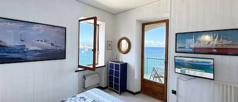 A bedroom with a bed, a window with a view of the sea, a door leading to a balcony, and a TV on the wall.
