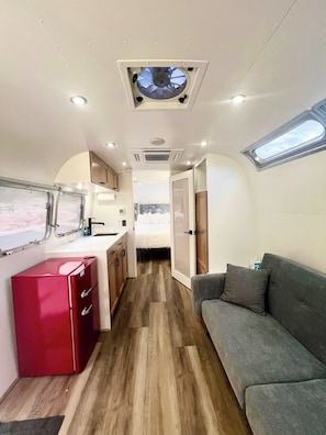 A compact RV interior with a kitchenette, a dining area, a gray sofa, and a small window.