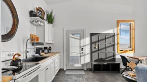 Une cuisine moderne avec des meubles blancs, une bouilloire noire et un plan de travail en bois. On y trouve une porte qui mène à une autre pièce et une fenêtre avec des stores.