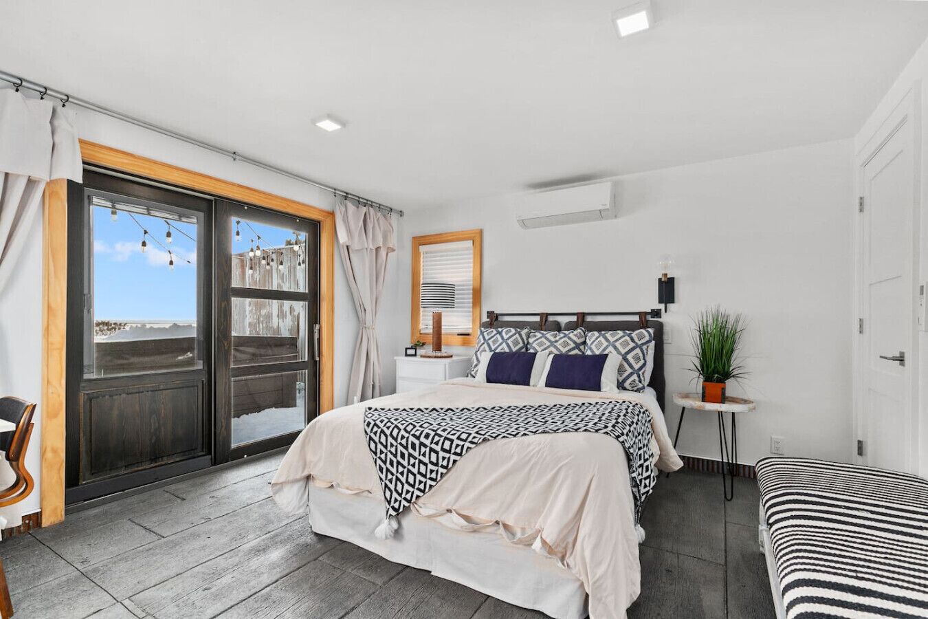 A bedroom with a bed, a bedside table, a chair, a small table with a plant, and a view of the sea through a sliding door.