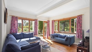 Uma sala de estar ampla com dois sofás, uma mesa de centro e um aparador para TV. Janelas grandes com cortinas oferecem luz natural e vista para o verde lá fora.