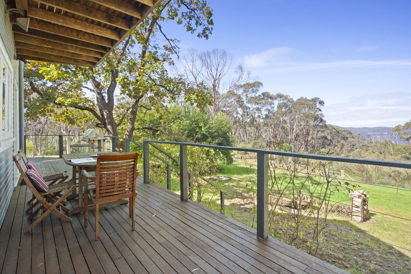Una terraza de madera con una mesa y sillas, con vistas a un paisaje cubierto de césped, árboles y una vista distante.