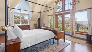 Quarto com cama de dossel, escrivaninha e cadeira, janelas amplas com cortinas, e vista para o exterior.