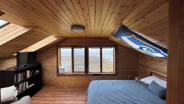 Una camera da letto accogliente in mansarda, con pareti in legno e soffitto spiovente. Dispone di una grande finestra con vista sul paesaggio, un letto con biancheria azzurra e una libreria con libri e una televisione.
