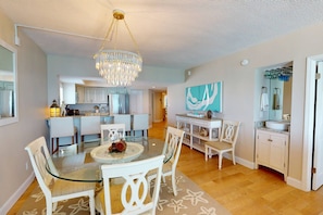 A modern kitchen with stainless steel appliances, a dining area with a glass table and white chairs, and a living area with a large window and a painting.