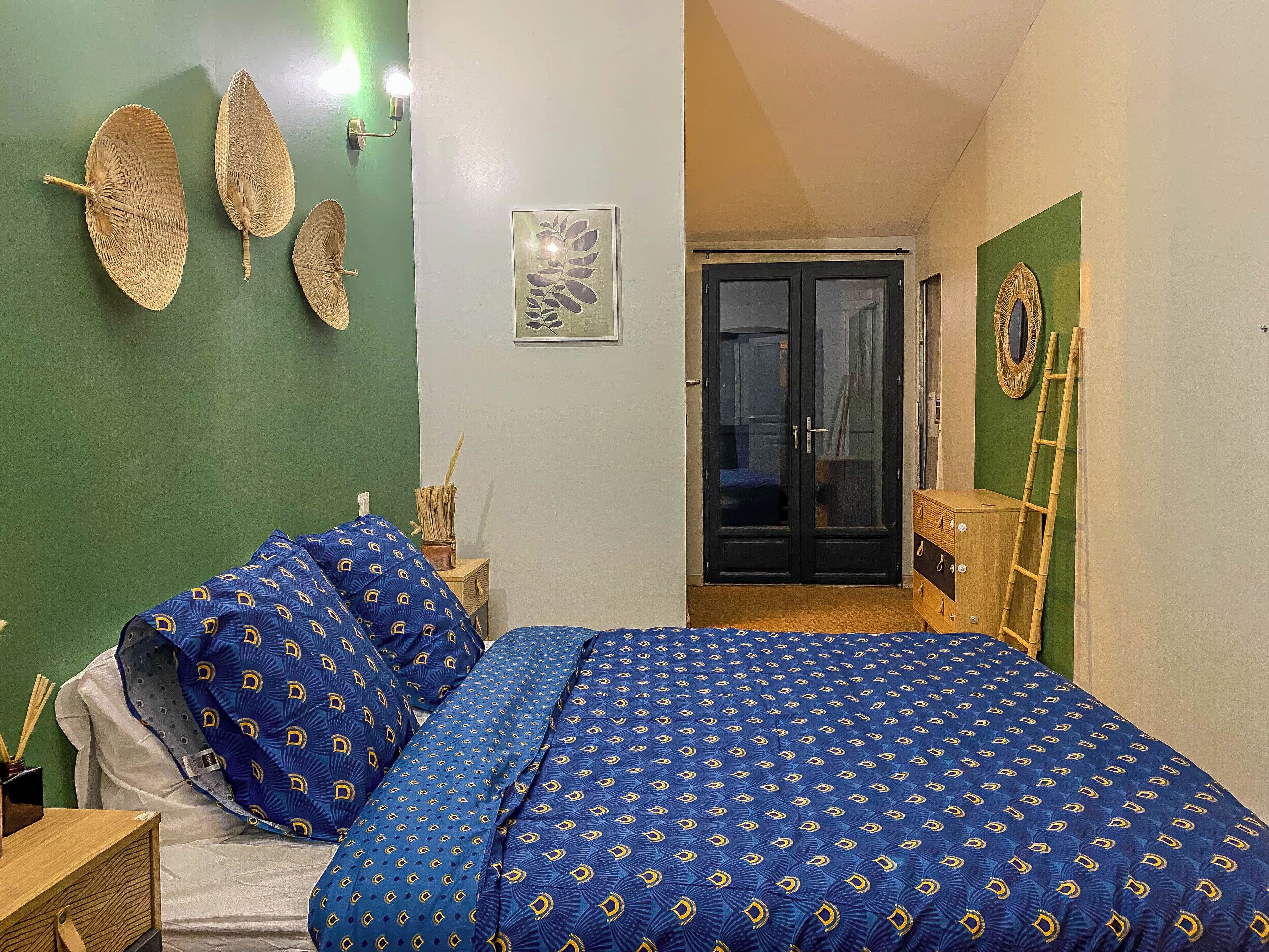 A bedroom with a bed, a nightstand, a lamp, a wall-mounted shelf, and a door leading to another room.