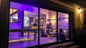 A modern interior with a large window, a sofa, a coffee table, and a bookshelf. - Carcalove - Pool, Billiards & Jacuzzi - 10pax (Fontiès-d'Aude)
