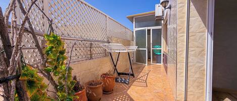 A balcony with potted plants, a drying rack, and a sliding glass door leading to a covered area.