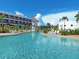 Una piscina de resort con sillas de descanso, un edificio con techo de paja y abundante vegetación.