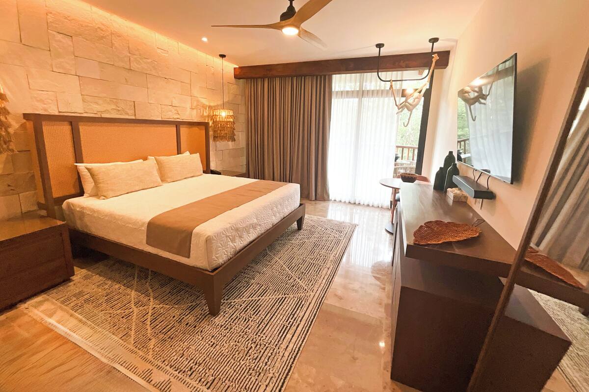A modern bedroom with a large bed, a ceiling fan, a stone wall, and a view of the outdoors.
