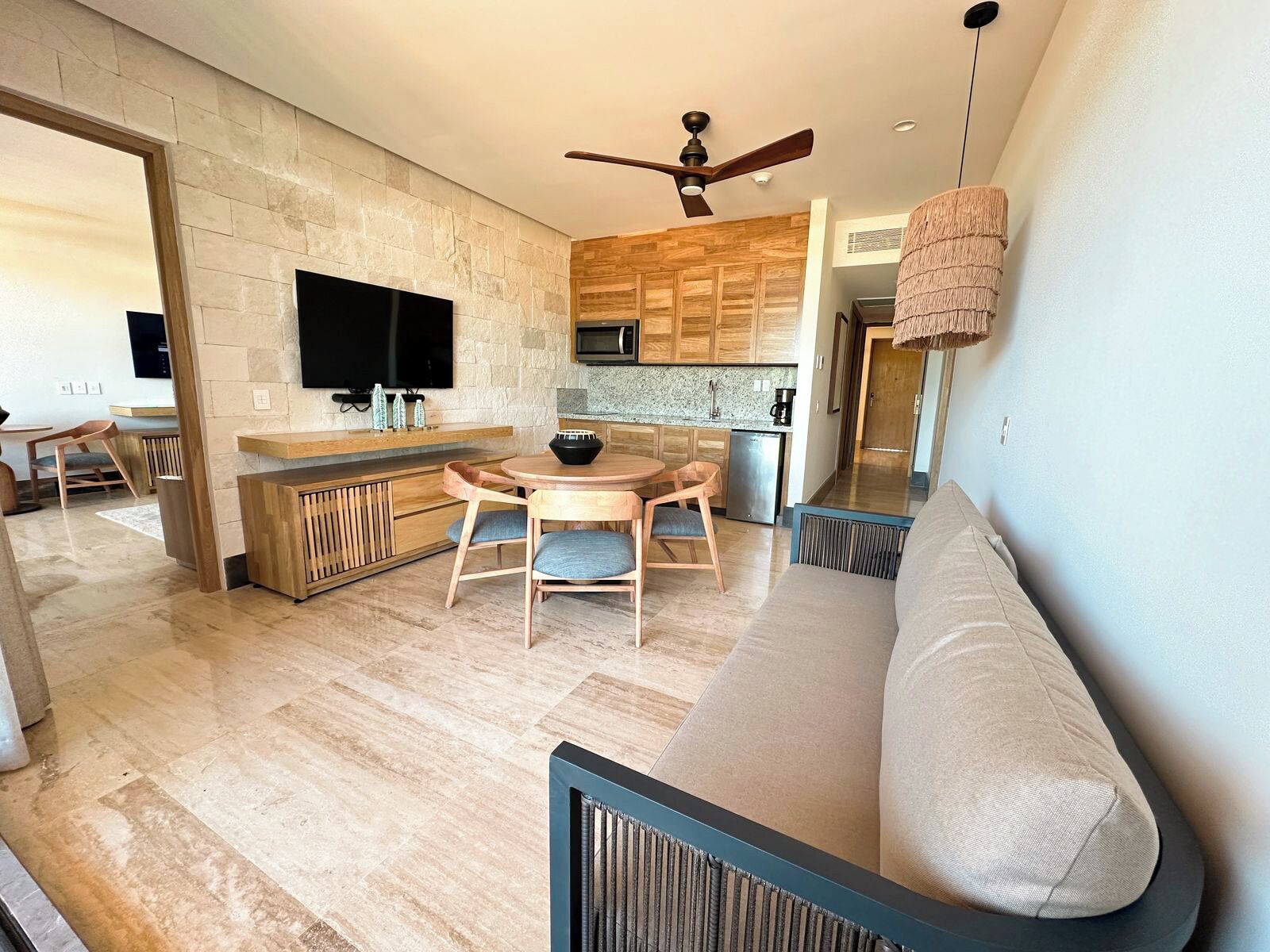 Un espace de vie moderne avec un mur en pierre, des meubles en bois, une cuisine avec des armoires en bois, un ventilateur de plafond et un téléviseur fixé au mur.