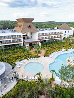 Un resort con una piscina grande, varias construcciones con techos de paja y abundante vegetación.