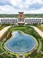 Un resort con una amplia piscina central rodeada de exuberante vegetación, un edificio de varios pisos con un techo de paja característico y vistas a colinas distantes.