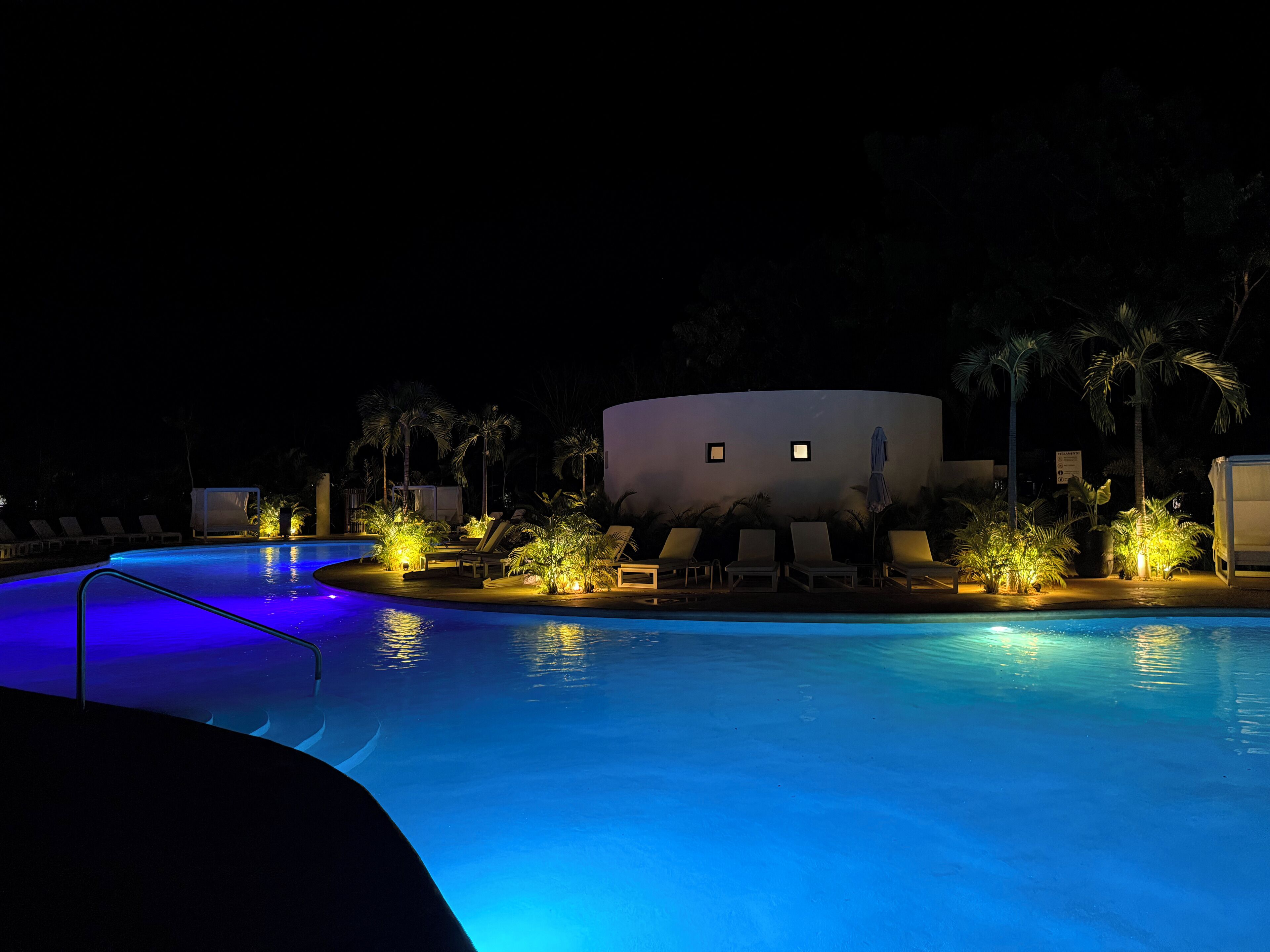 A nighttime swimming pool area with lounge chairs and palm trees, illuminated by ambient lighting.