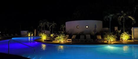 A nighttime swimming pool area with lounge chairs and palm trees, illuminated by ambient lighting.