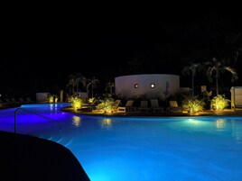 Un área de piscina iluminada con luces ambientales, sillones de descanso y palmeras.