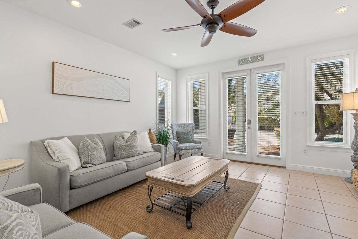 A living room with a grey sofa, a wooden coffee table, a ceiling fan, and large windows with blinds.