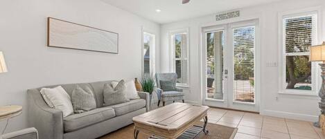 A living room with a grey sofa, a wooden coffee table, a ceiling fan, and large windows with blinds.