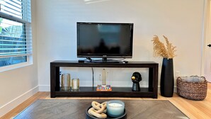Una sala de estar moderna con un televisor de pantalla plana sobre un mueble oscuro, un jarrón decorativo con plantas secas y una mesita con velas y aperitivos.