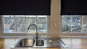 Une cuisine équipée d’un évier en acier inoxydable, d’un robinet et d’un égouttoir à vaisselle. Une fenêtre offre une vue sur un quartier résidentiel.