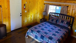 A wooden cabin bedroom with a bed, a nightstand, a window with curtains, and a door.
