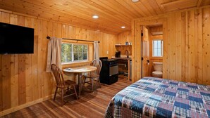 Interior de una cabaña de madera con una cama, un escritorio, una silla, un televisor y una ventana.