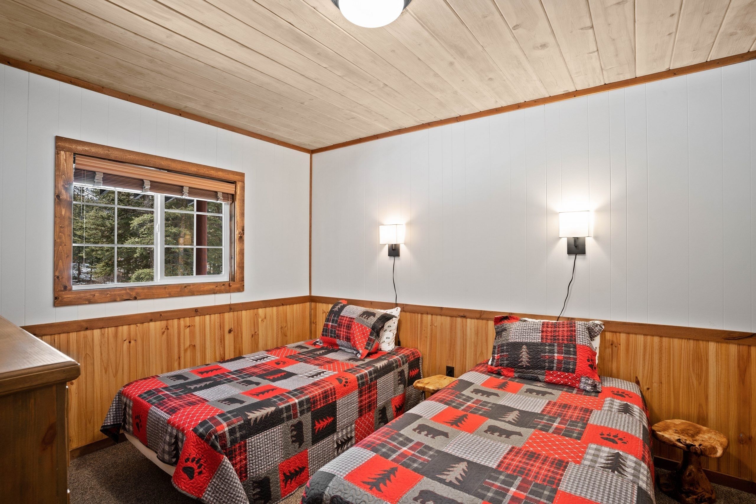 A cabin-style bedroom with two beds, wooden walls, a window with blinds, and wall-mounted lights.