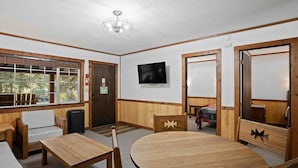 Una habitación con una mesa redonda de madera, dos sillas de madera, un televisor montado en la pared y una ventana con vistas a árboles.