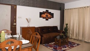 A living room with a brown sofa, a dining table with chairs, a ceiling fan, and a wall-mounted artwork. - Tulsi Home Stay (Mumbai)