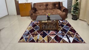 A living room with a brown sofa, a coffee table, a patterned rug, a wall-mounted artwork, a potted plant, and a wooden cabinet. - Tulsi Home Stay (Mumbai)