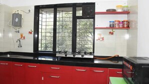 A kitchen with red cabinets, a gas stove, a window with a view of trees, and a water heater on the wall. - Tulsi Home Stay (Mumbai)