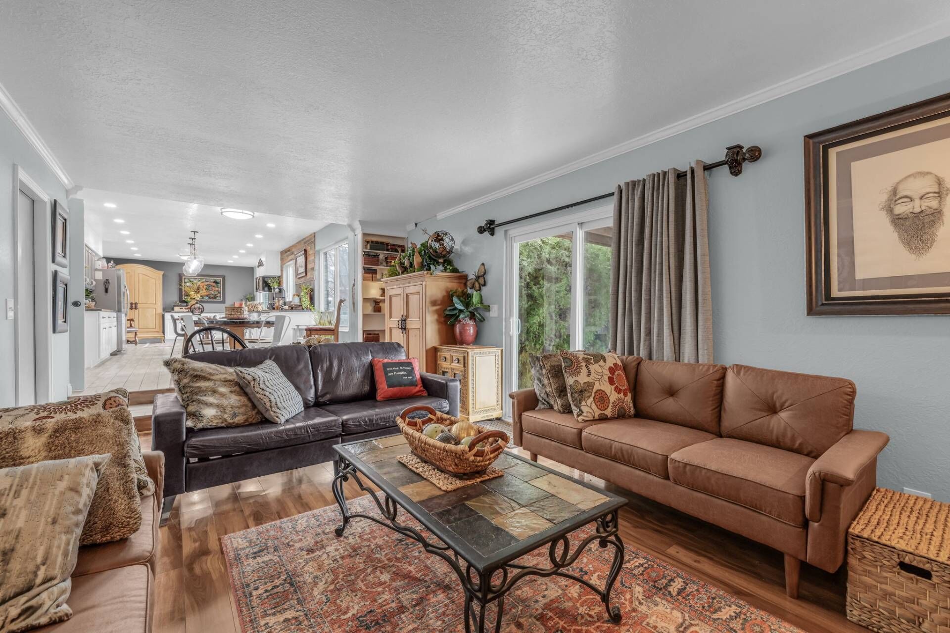 A spacious living room with a brown leather sofa, a black leather sofa, and a patterned rug. There is a dining area in the background with a wooden table and chairs.