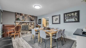 A dining area with a stone fireplace, a wooden dining table set for four, and a modern kitchen in the background.