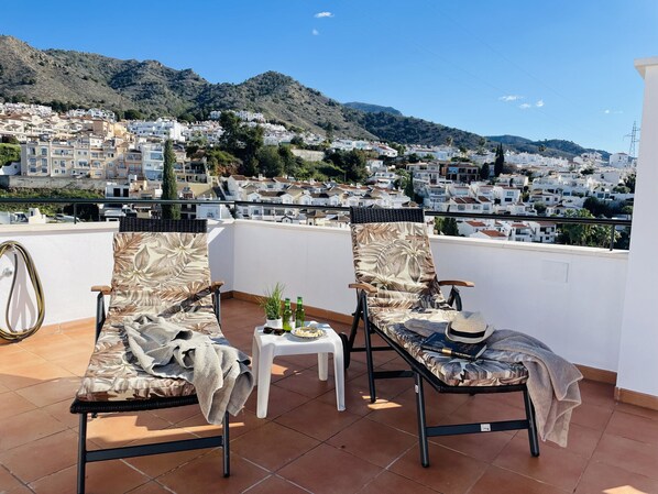 A terrace with two lounge chairs, a small table, and a view of a hillside town.