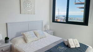 A neatly arranged bedroom with a bed, pillows, a bedside table, a lamp, and a window with a view of a coastal area. - Apartment Mar de Nerja Sunset (Nerja)