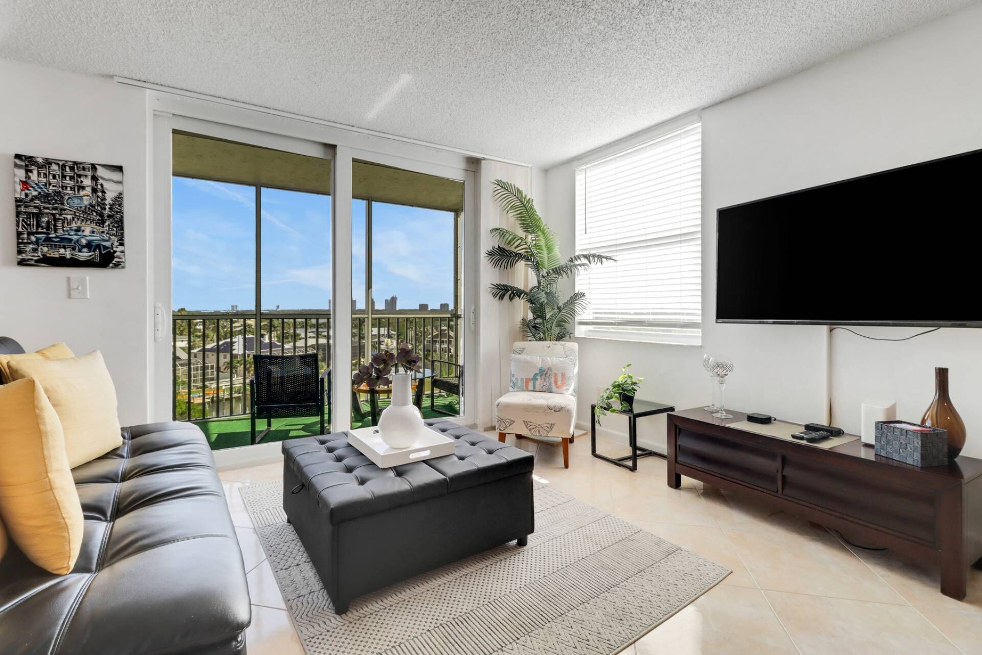 A modern living room with a grey sofa, a black ottoman, a TV, and a balcony with a view.