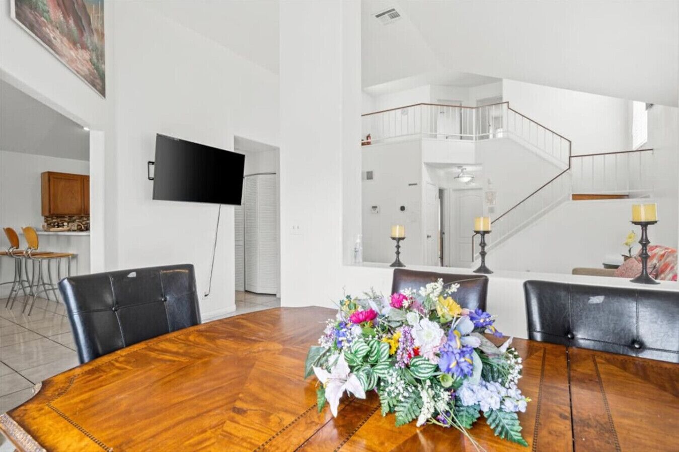 A dining area with a wooden table, chairs, and a floral centerpiece. A flat-screen TV is mounted on the wall, and a staircase is visible in the background.