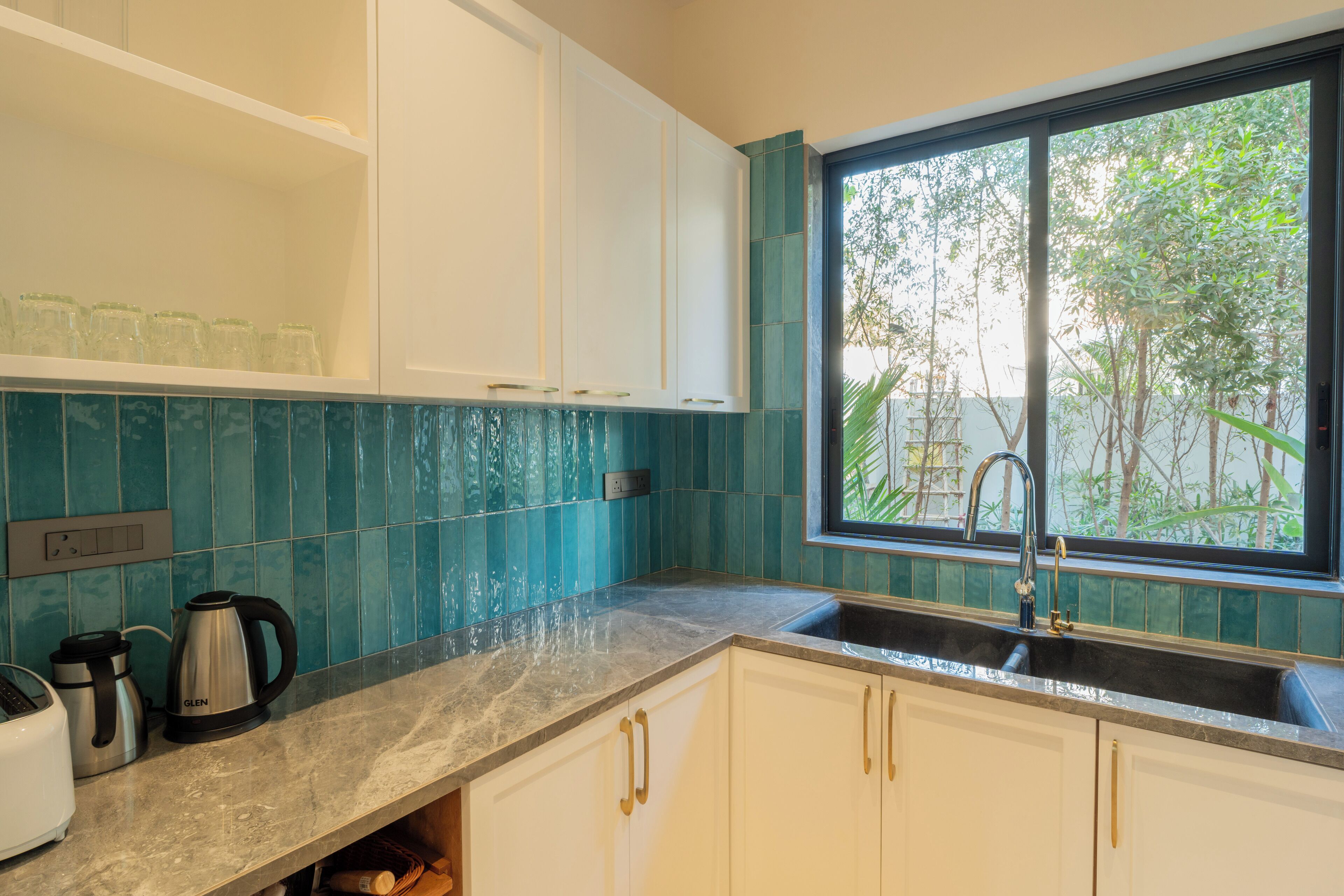A modern kitchen with white cabinets, a blue-tiled backsplash, a stainless steel coffee maker, and a window with a view of greenery.