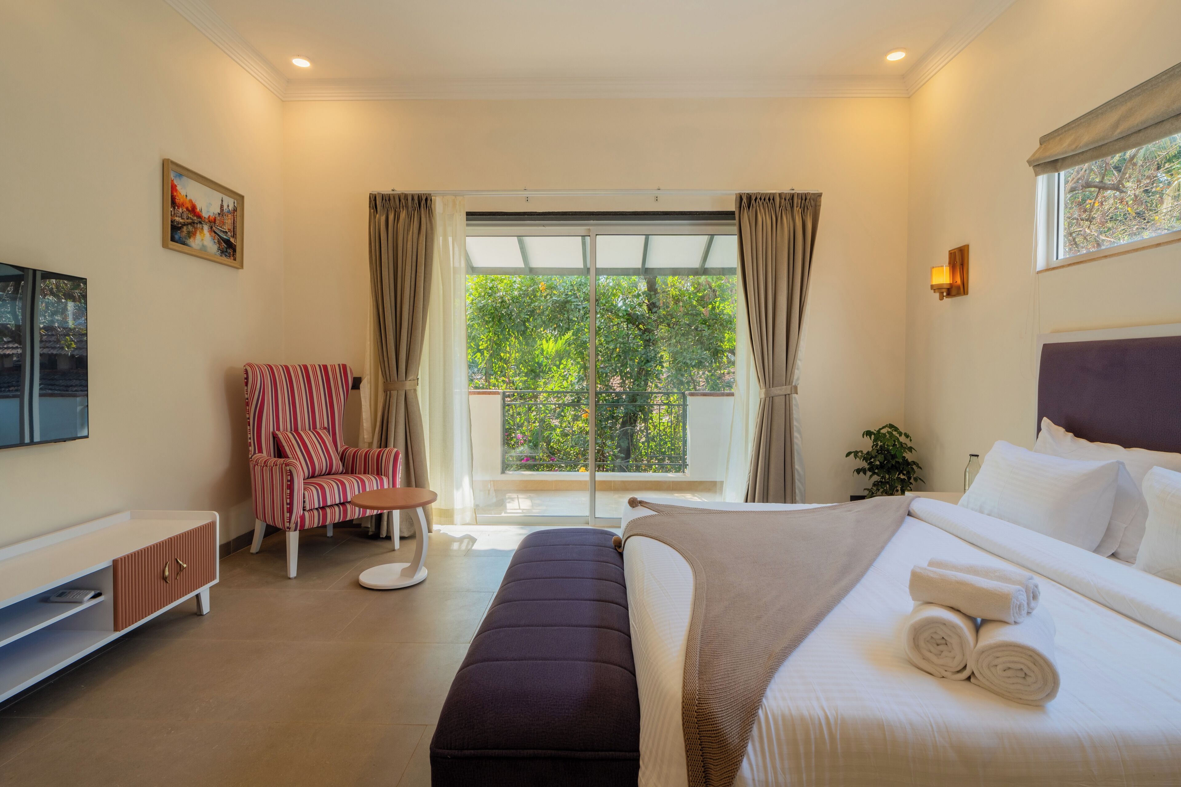 A hotel room with a bed, a chair, a TV, and a balcony with a view of greenery.